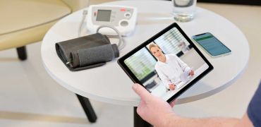 Patient sitzt am Tisch mit Tablet, Ärztin führt Telekonsultation durch.