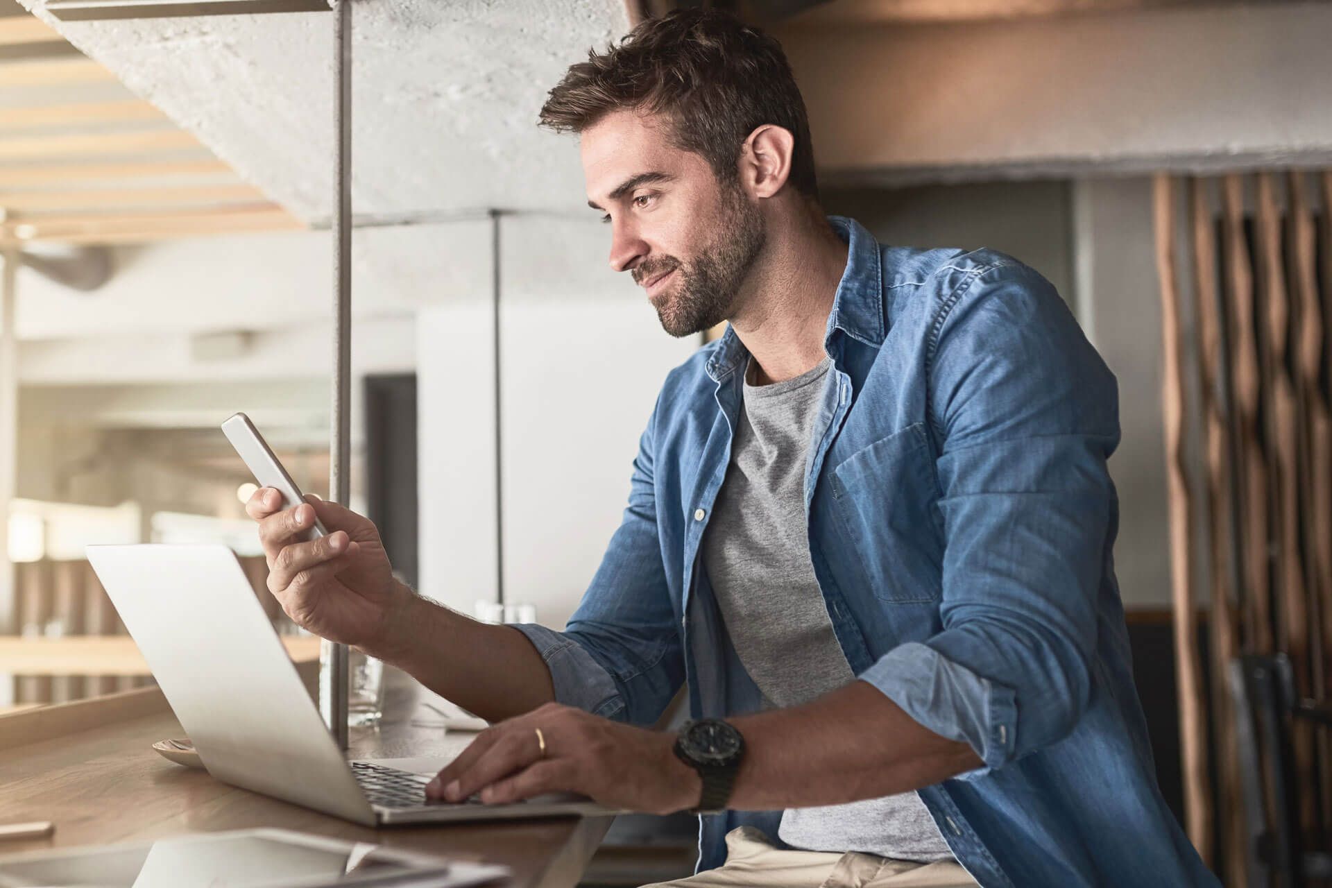 Junger Mann sitzt vor seinem Laptop und hält sein Handy in der Hand.