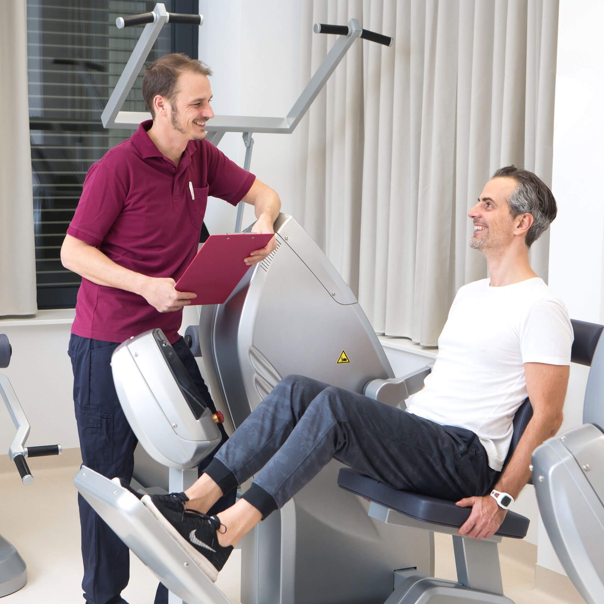 Patient trainiert am Kraftgerät, Physiotherapeut gibt Übungsanweisungen.